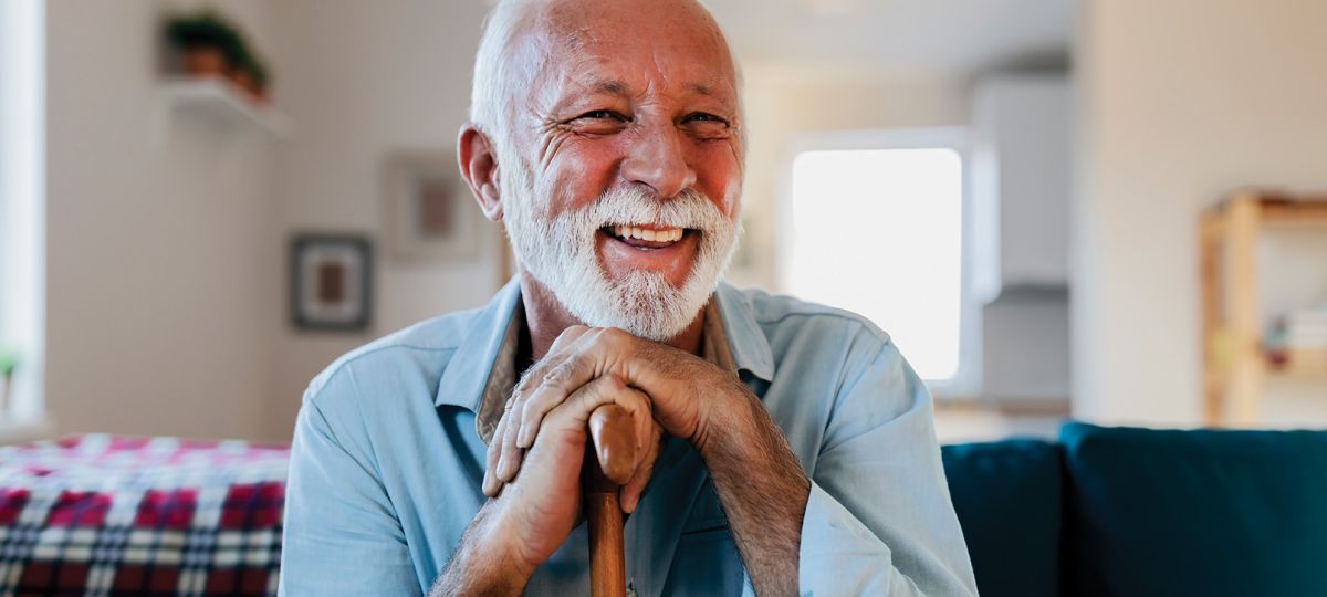 Beautiful bearded smiling older man holding a cane while sitting on a couch.