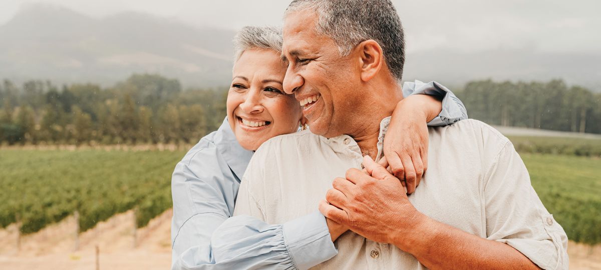 Older Hispanic couple, with the woman hugging the man, standing in front of a scenic outdoor backdrop with mountains and a vineyard.